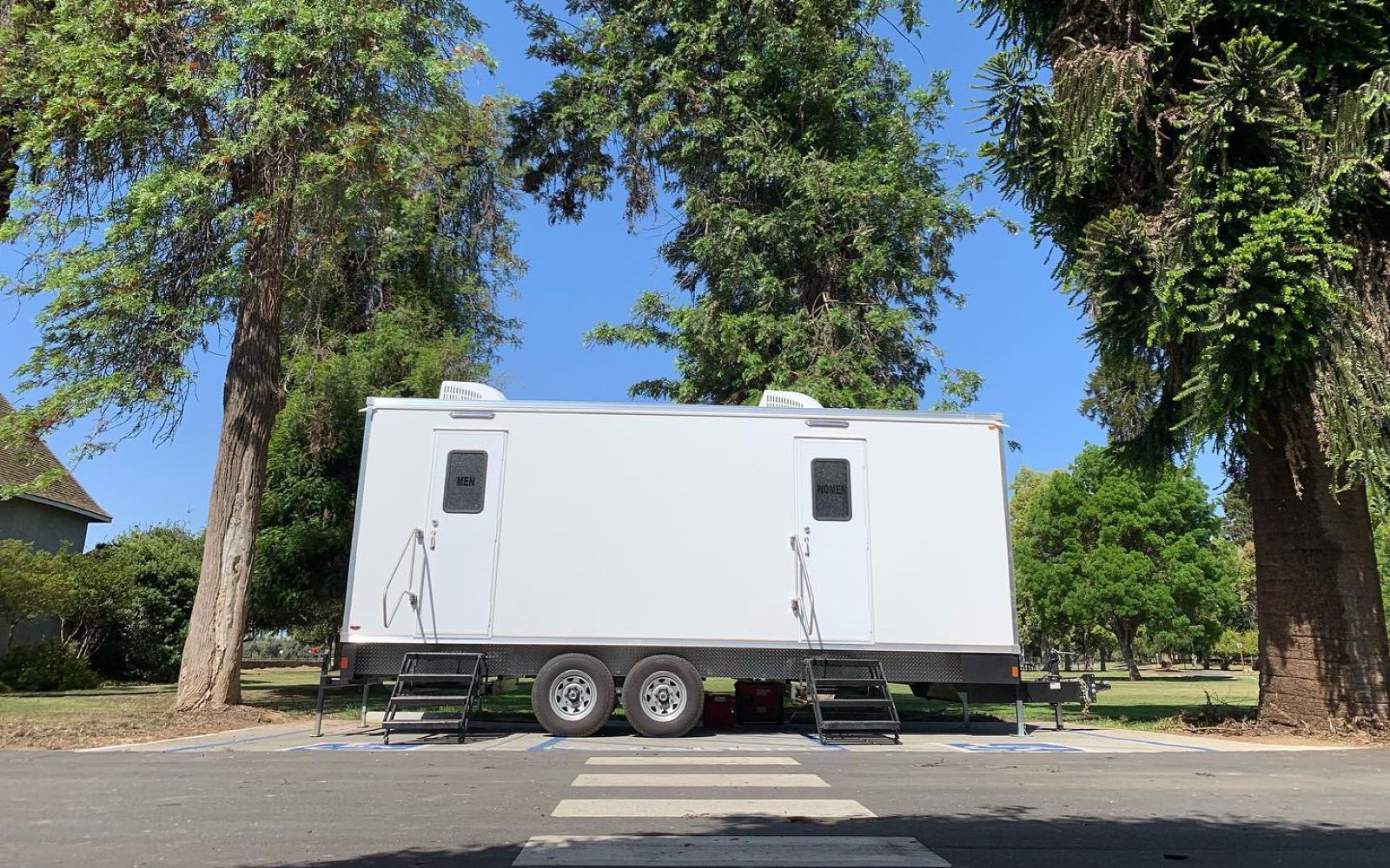9-stall bathroom trailer with two entrances parked under tall trees beside a city park, The Lavatory NorCal