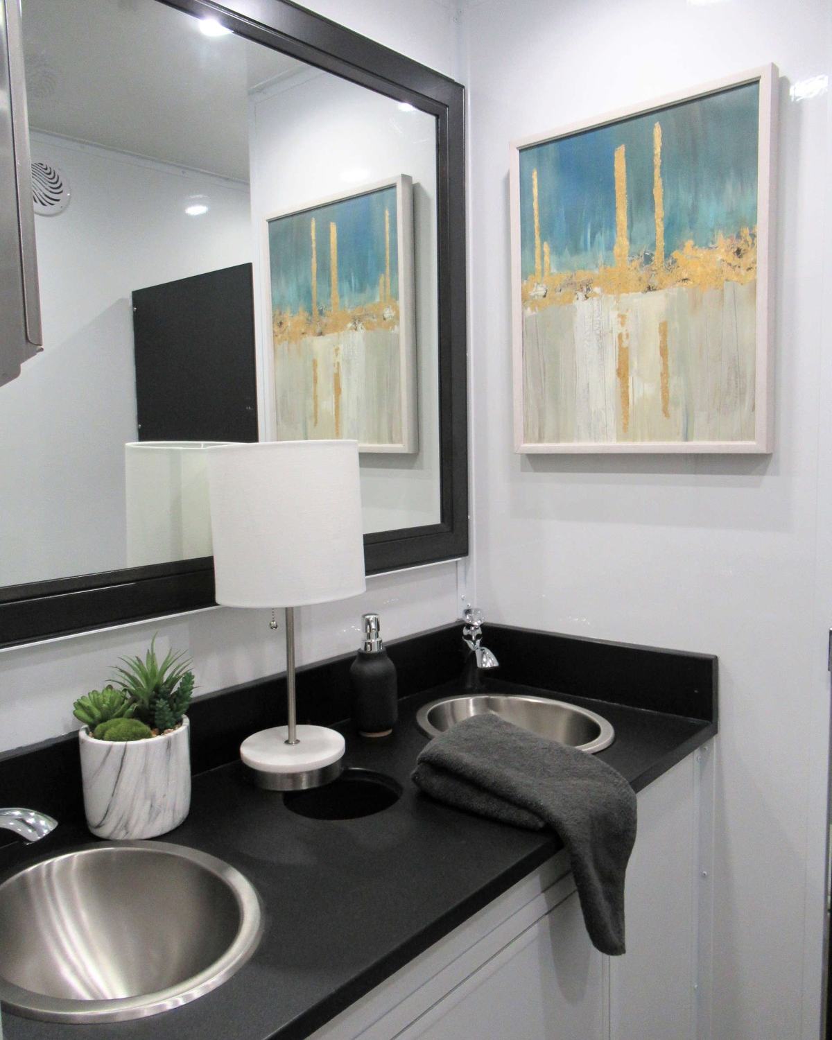 Double vanity with stainless sinks, a lamp, a framed mirror and framed art inside a luxury bathroom trailer, The Lavatory NorCal