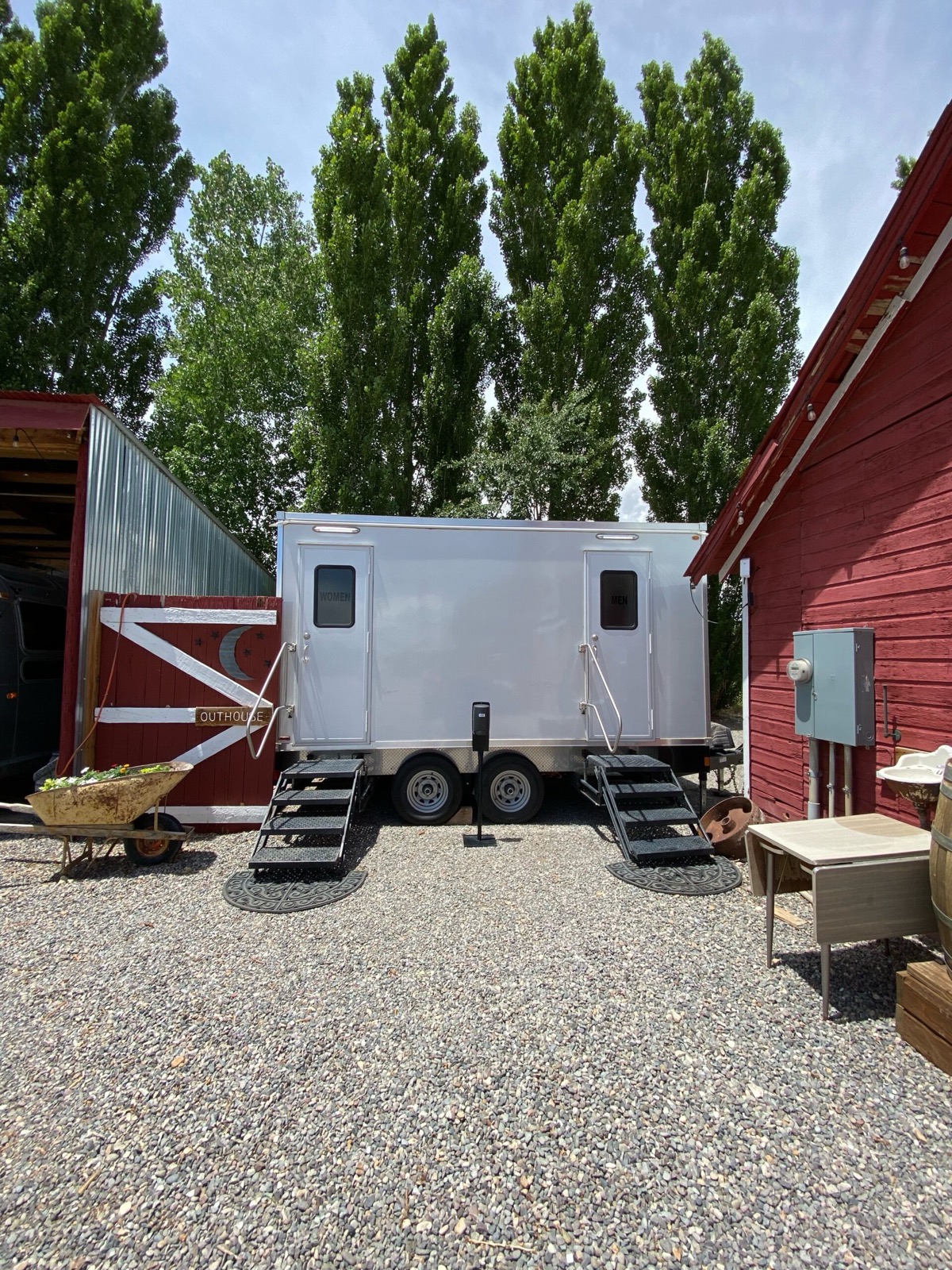 Two-door bathroom trailer set on gravel between a red barn and a shed at a ranch property, The Lavatory NorCal
