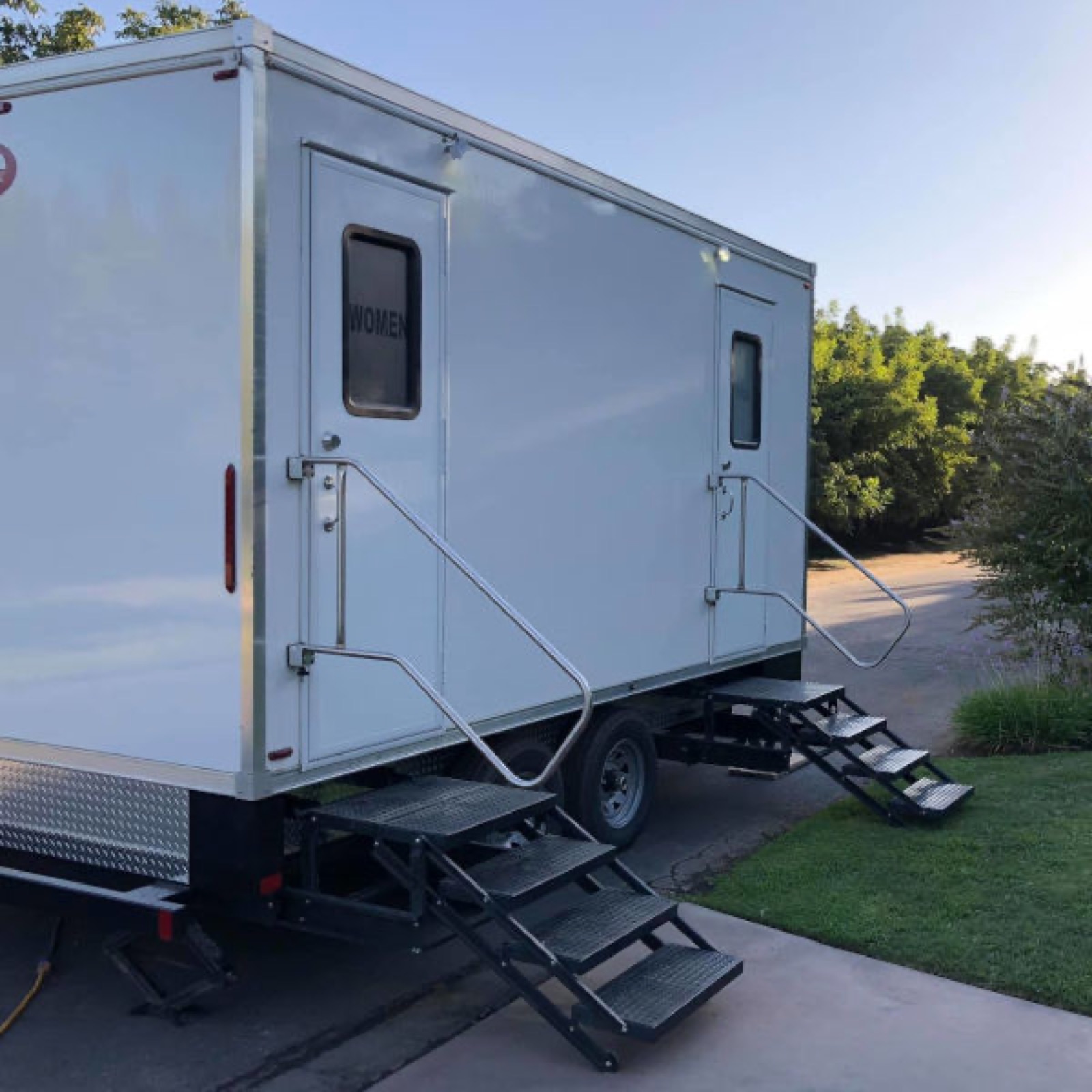 Side view of a white restroom trailer with folding steel stairs and railings set on a paved driveway, The Lavatory NorCal