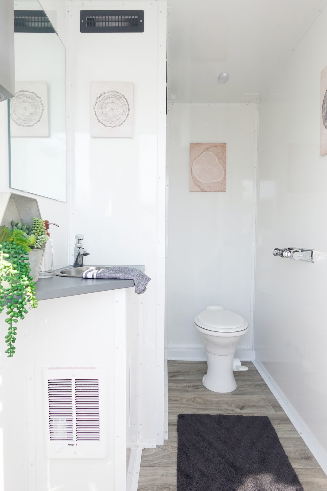 Inside a 2-stall bathroom trailer room: vanity with sink and succulents, framed art, toilet and wood-look floor, The Lavatory NorCal