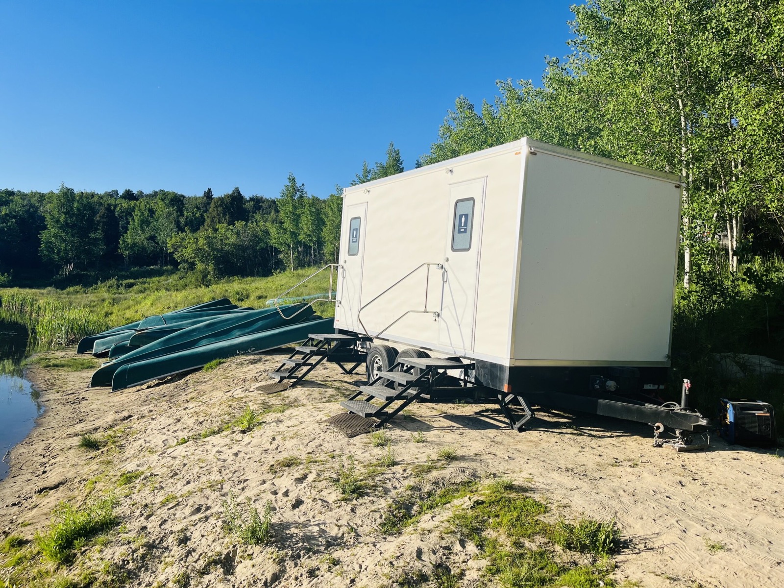 Two-stall portable restroom trailer on a sandy lake shore beside stacked canoes, The Lavatory NorCal