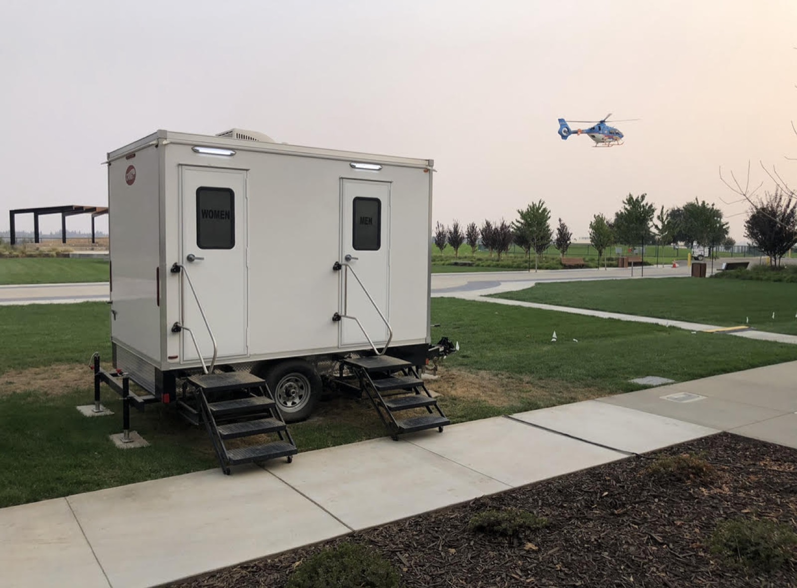 Temporary 2-stall restroom trailer on a park lawn in Napa during a smoky wildfire week with a medical helicopter overhead, The Lavatory NorCal