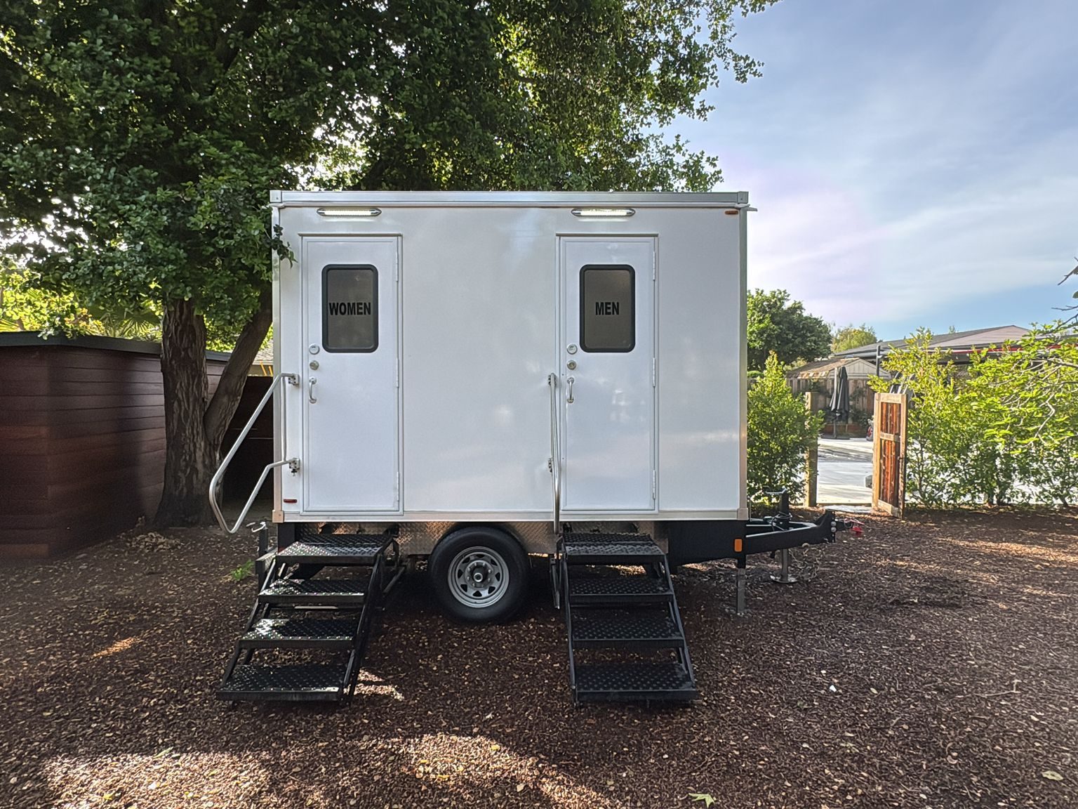 2-stall bathroom trailer with women's and men's doors and folding steps in a shaded backyard, The Lavatory NorCal