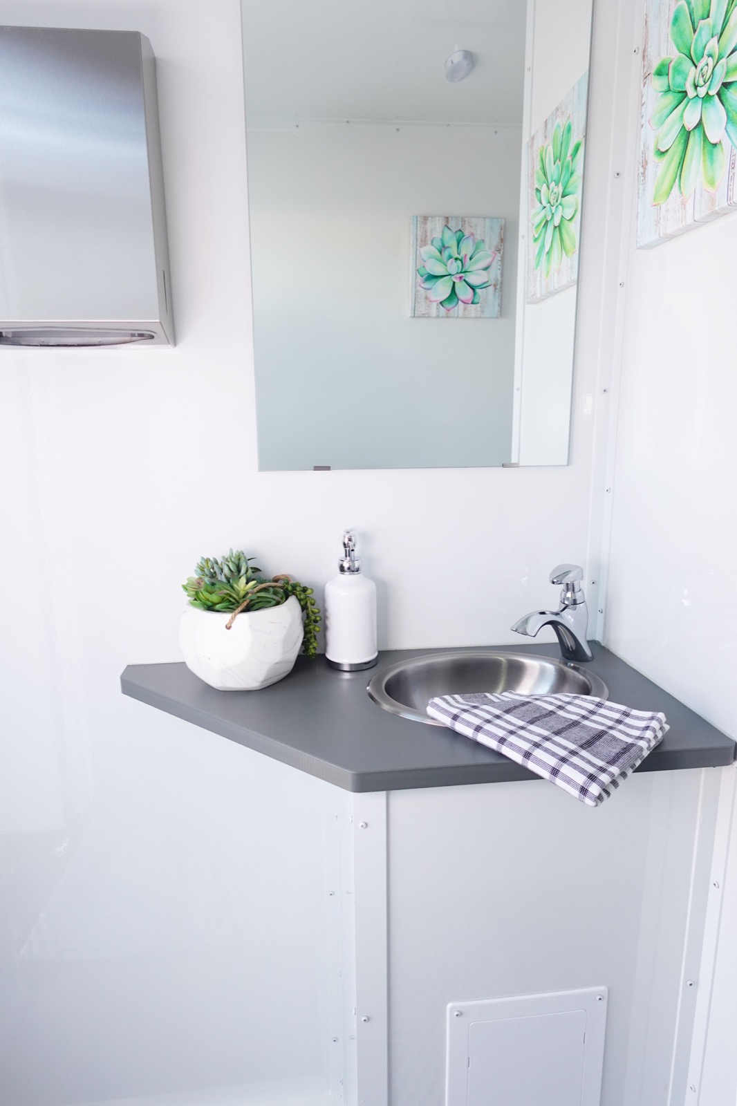 Corner sink vanity with a framed mirror and succulent art inside a 3-stall luxury bathroom trailer, The Lavatory NorCal