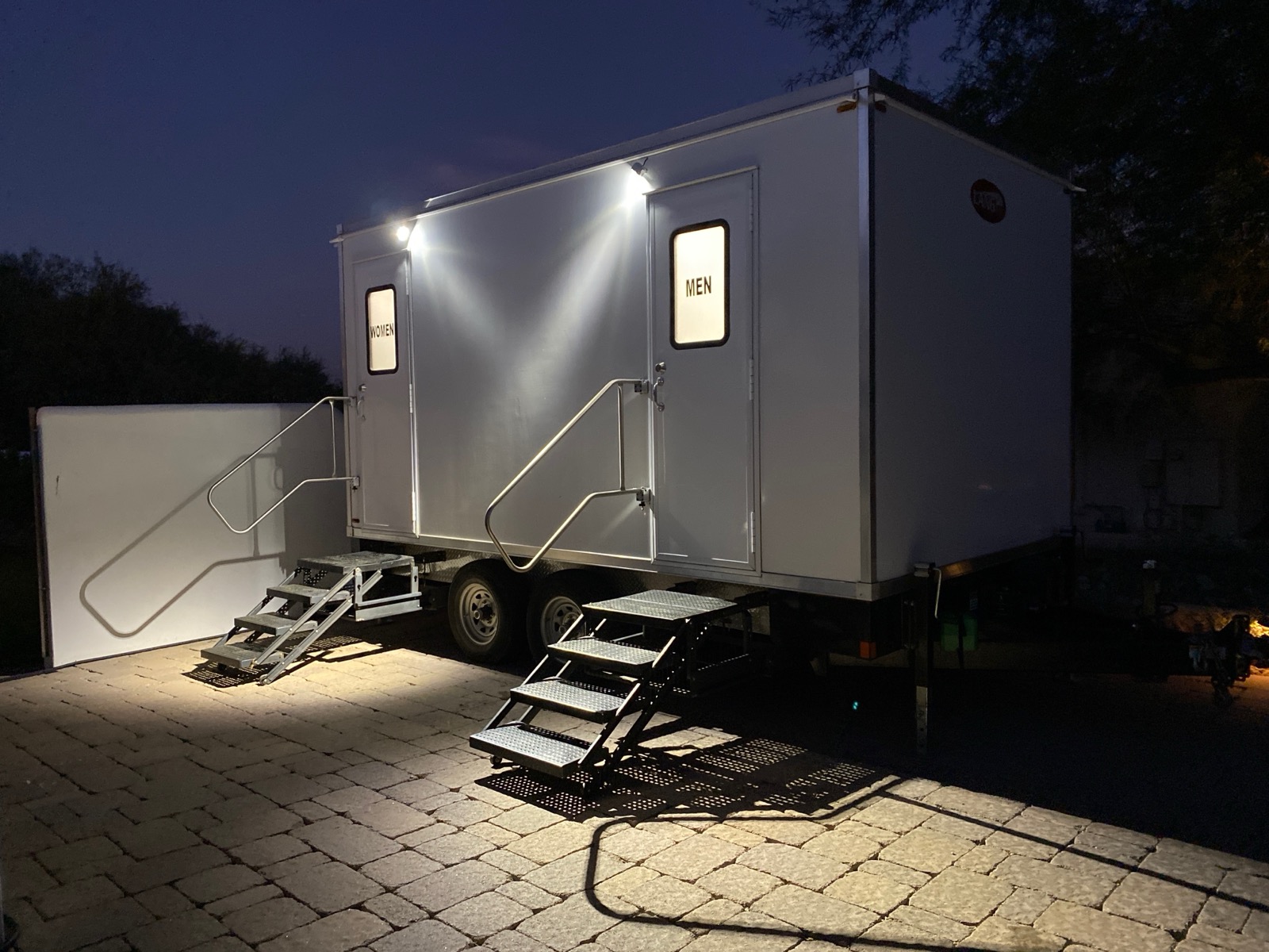 5-stall bathroom trailer with lit women's and men's entrances and folding stairs on a stone patio at night, The Lavatory NorCal