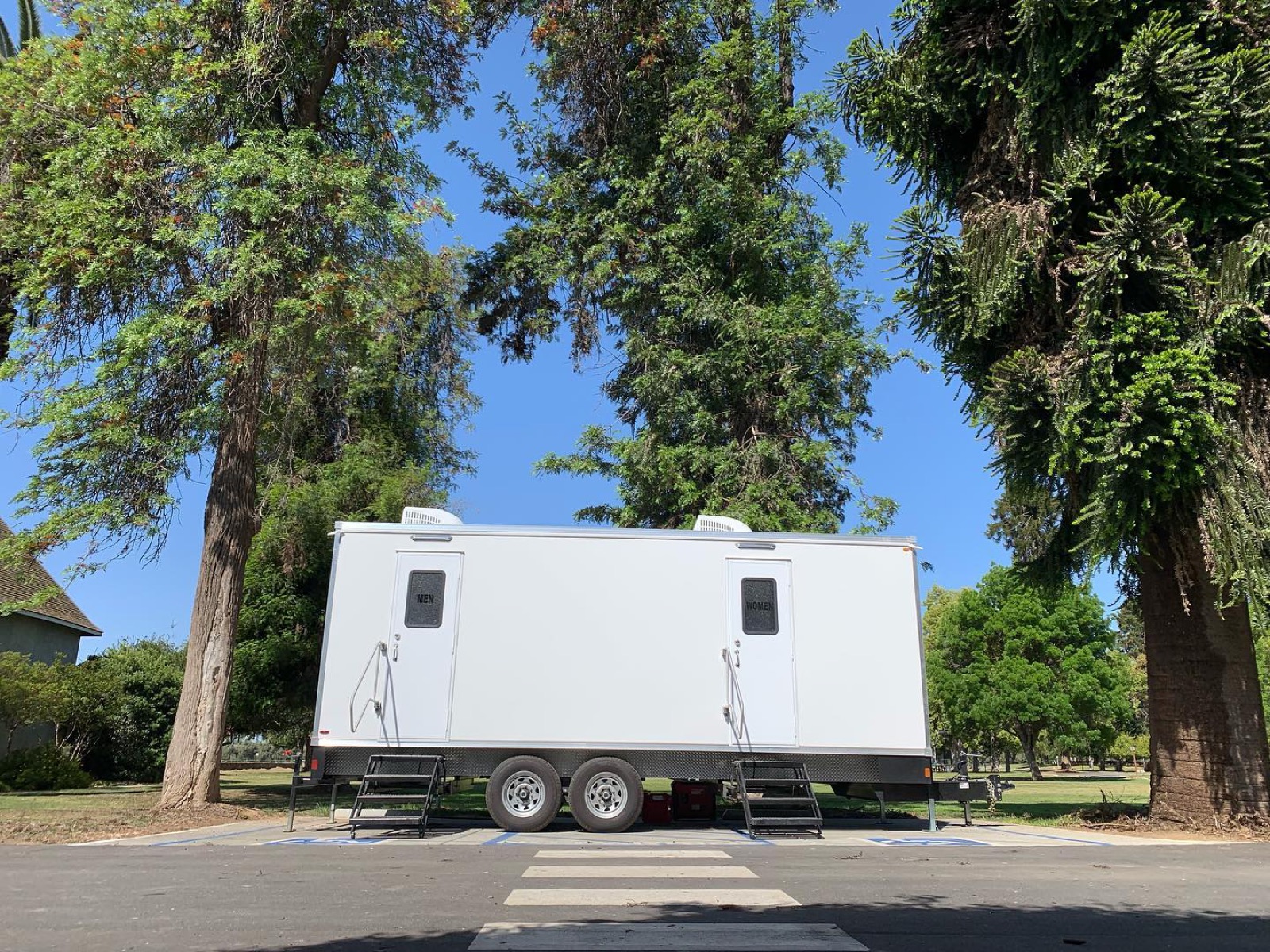 9-stall restroom trailer parked curbside under tall trees beside a city park, The Lavatory NorCal