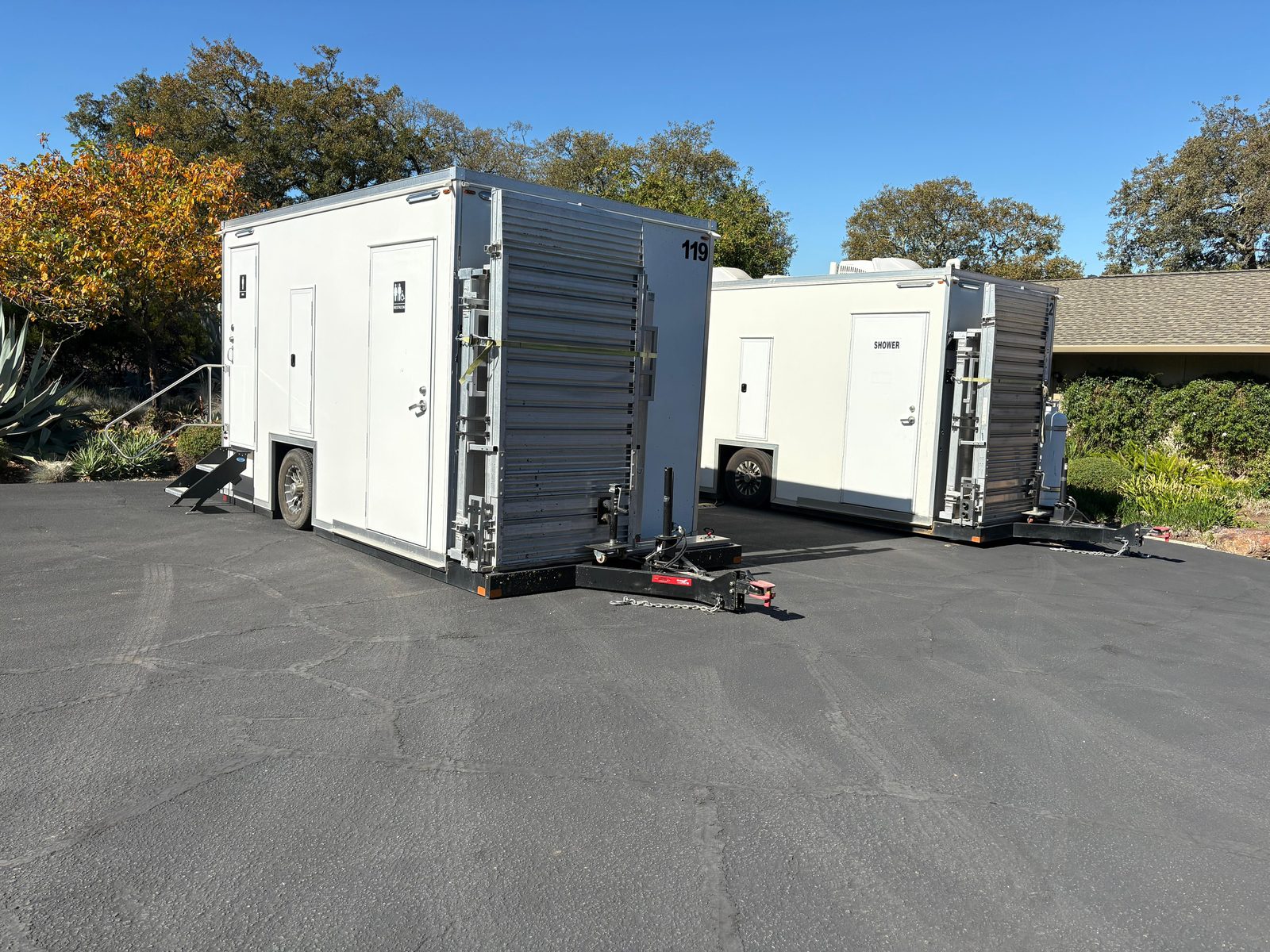 ADA restroom trailer and ADA shower unit parked on a lot lined with oak trees, ramp systems folded against the hitch end, The Lavatory NorCal