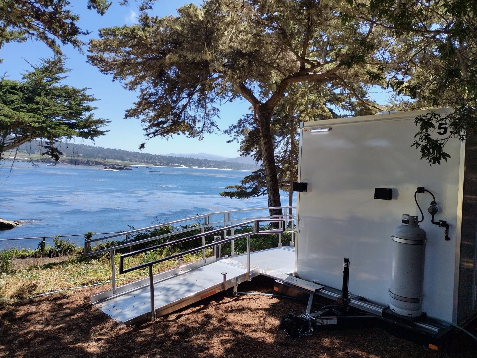 Accessible restroom trailer with a railed ramp under cypress trees on a windy coastal bluff, The Lavatory NorCal
