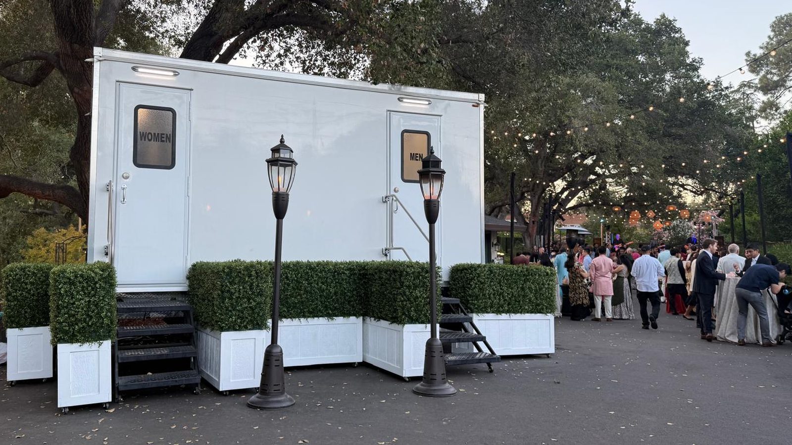 Restroom trailer with women's and men's doors dressed with boxwood planters and lantern posts at an evening garden party under oak trees, The Lavatory NorCal