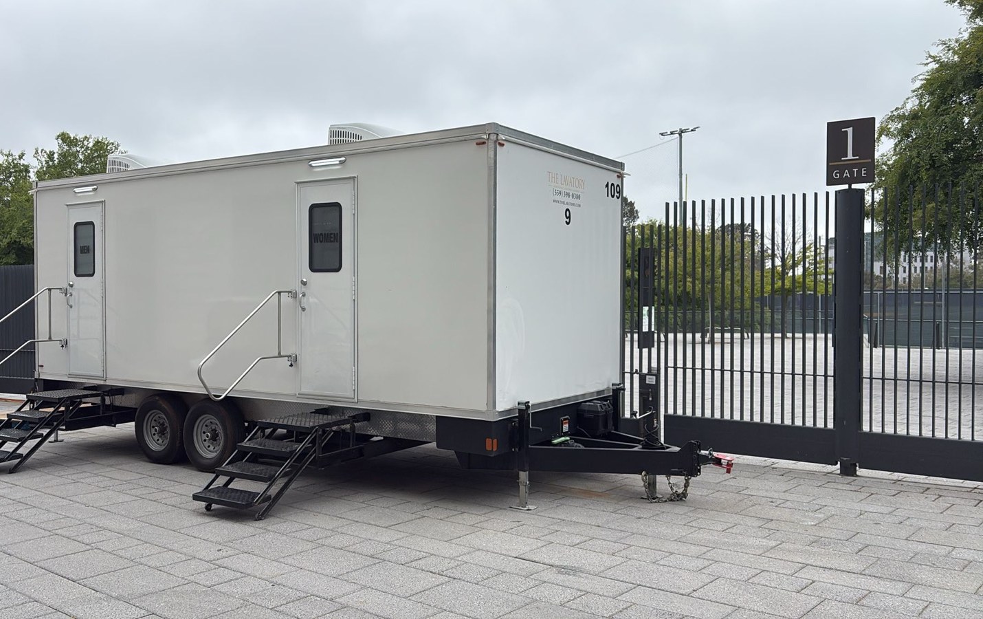 9-stall luxury bathroom trailer with men's and women's doors and steel stairs set on pavers beside a stadium gate in Berkeley, The Lavatory NorCal