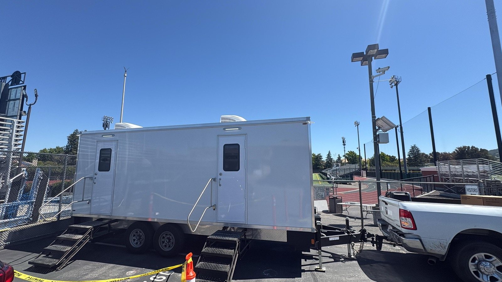 Portable bathroom trailer with women's and men's doors and railed stairs parked beside a ball field in Concord, The Lavatory NorCal