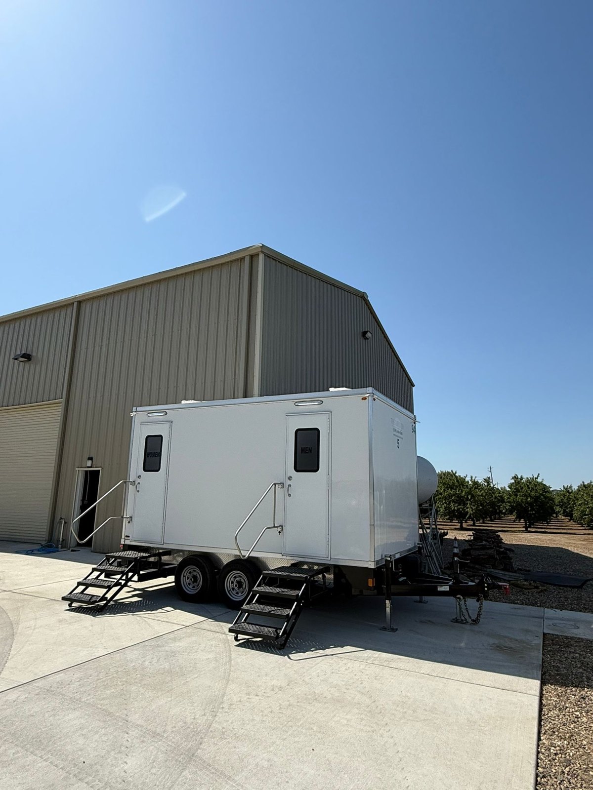 5-stall bathroom trailer parked on a concrete pad beside a metal farm building and an orchard south of Elk Grove, The Lavatory NorCal