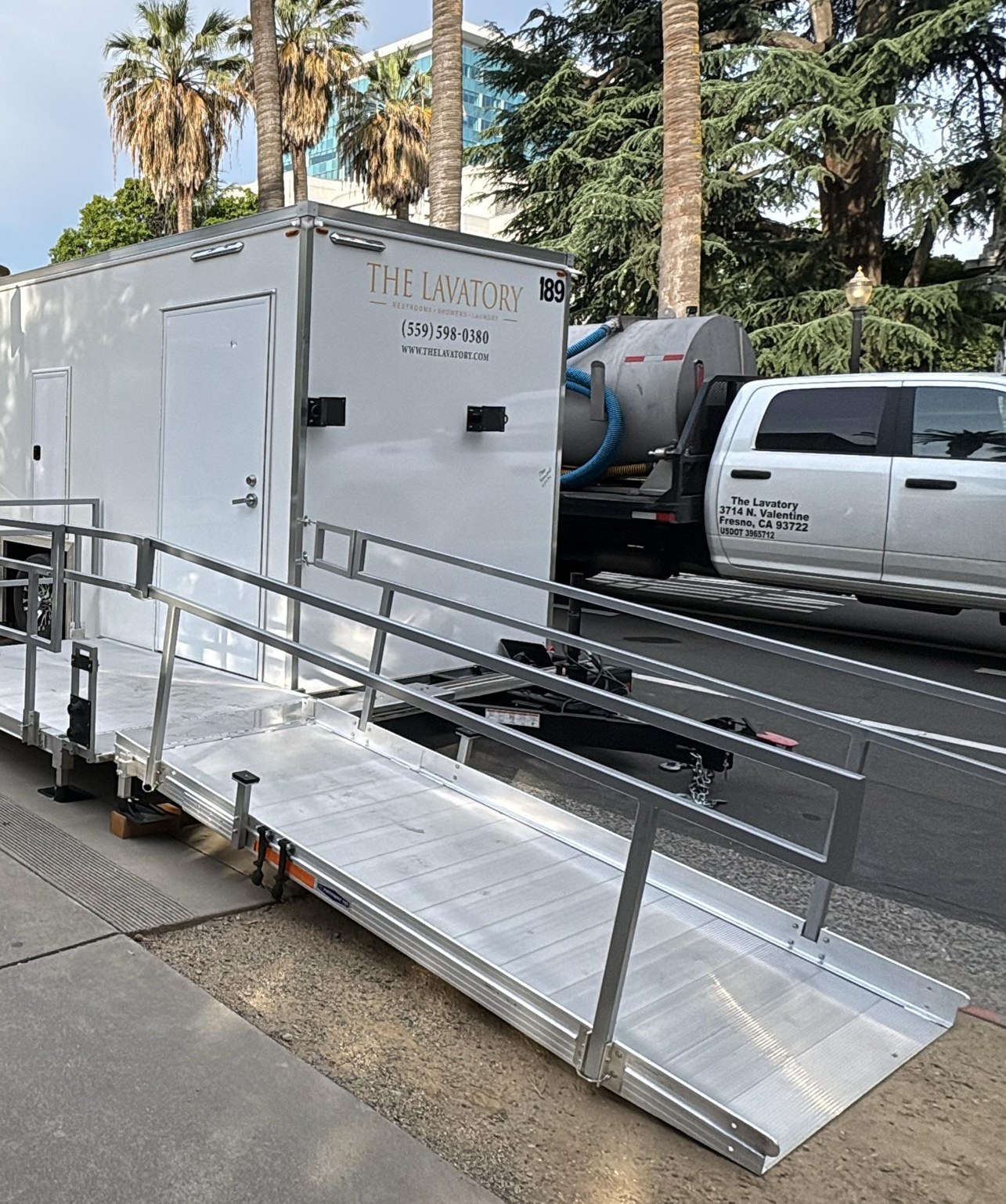 Accessible restroom trailer with a long aluminum ramp set along a downtown Sacramento sidewalk, with our service truck at the curb, The Lavatory NorCal