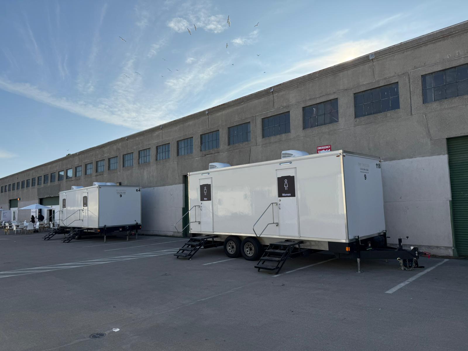Row of 9-stall portable restroom trailers lined up along a long concrete warehouse in San Francisco for a corporate keynote event, The Lavatory NorCal