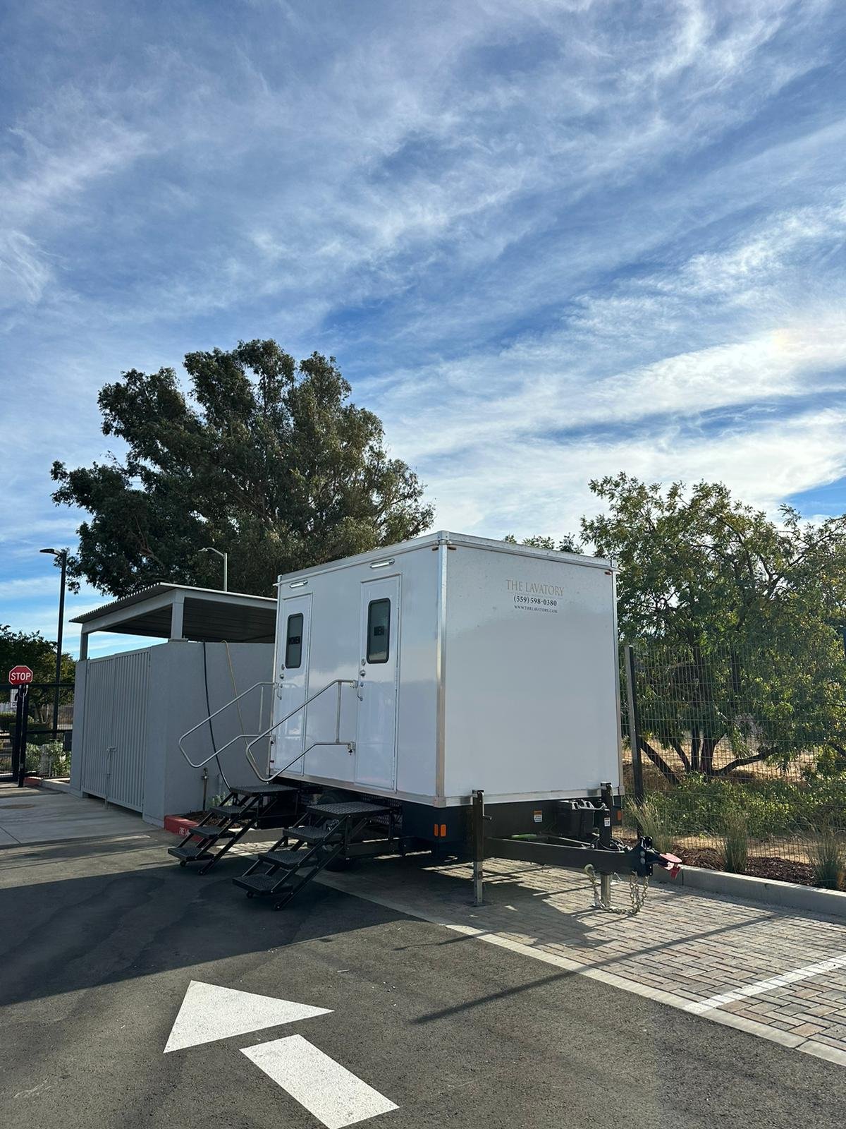 2-stall restroom trailer with two entry stairs parked beside a paved campus lot under a big shade tree in the South Bay, The Lavatory NorCal
