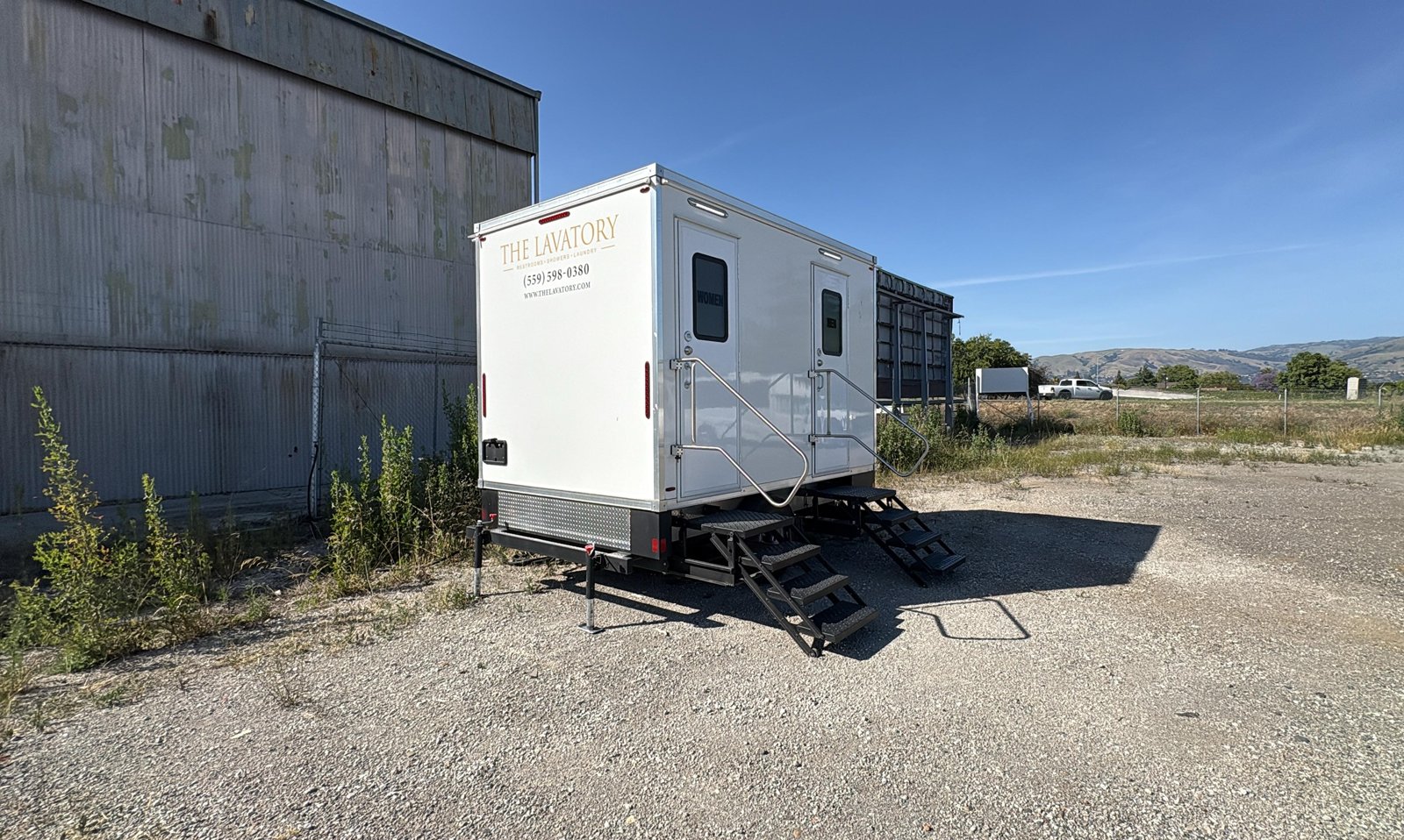 2-stall restroom trailer on a gravel yard beside a metal warehouse with the South Bay foothills behind, The Lavatory NorCal