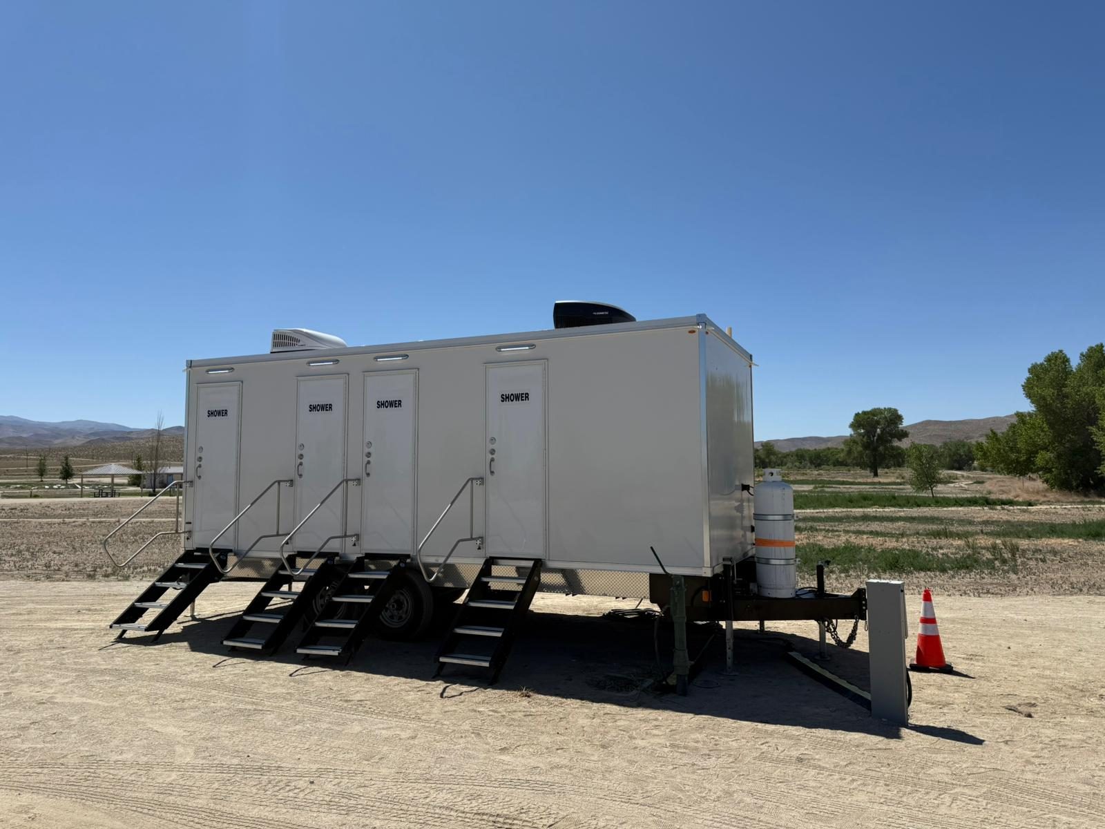 Shower trailer with four shower doors and folding stairs on a gravel lot at a park with cottonwood trees, The Lavatory NorCal
