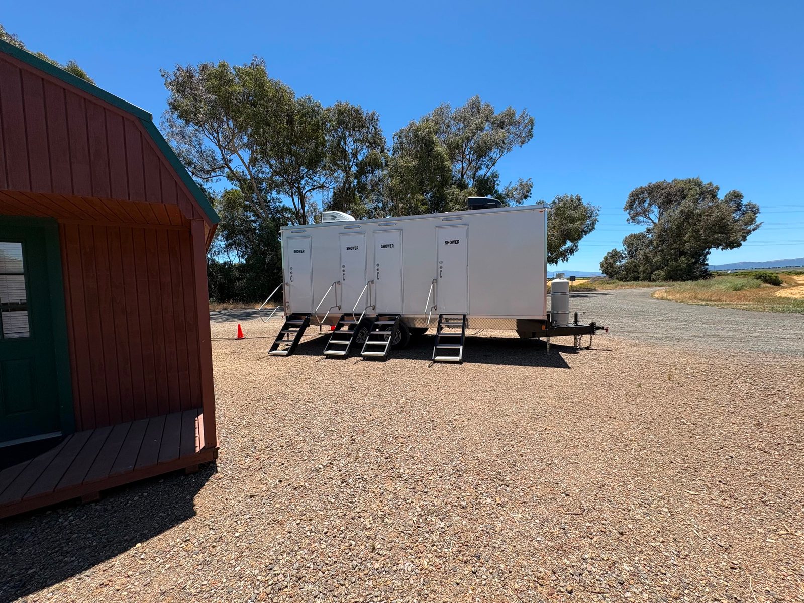 Shower trailer with four shower doors and folding stairs on a wide gravel lot beside a small barn-style shed, The Lavatory NorCal