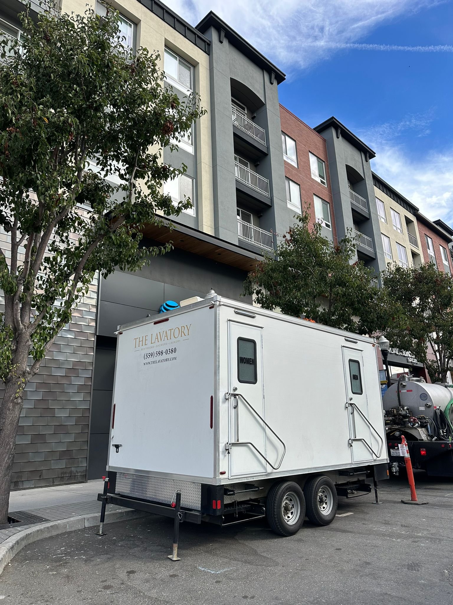 Bathroom trailer with women's and men's doors parked beside new apartments in Sunnyvale with our service truck behind it, The Lavatory NorCal